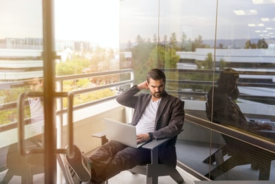 白天，一名男子坐在灰色扶手椅上，在大楼阳台上使用银色笔记本电脑